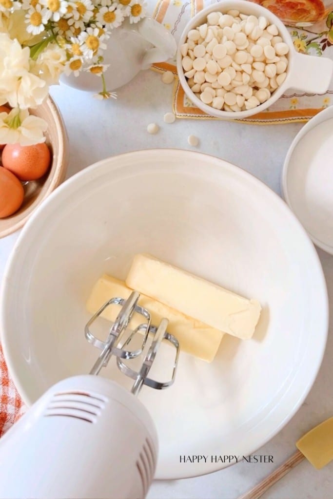 A mixing bowl with two sticks of butter and an electric hand mixer. Surrounding the bowl on a countertop are white chocolate chips in a bowl, two eggs, flowers, a cup of sugar, and a folded gingham cloth. A recipe card peeks out from under the ingredients.