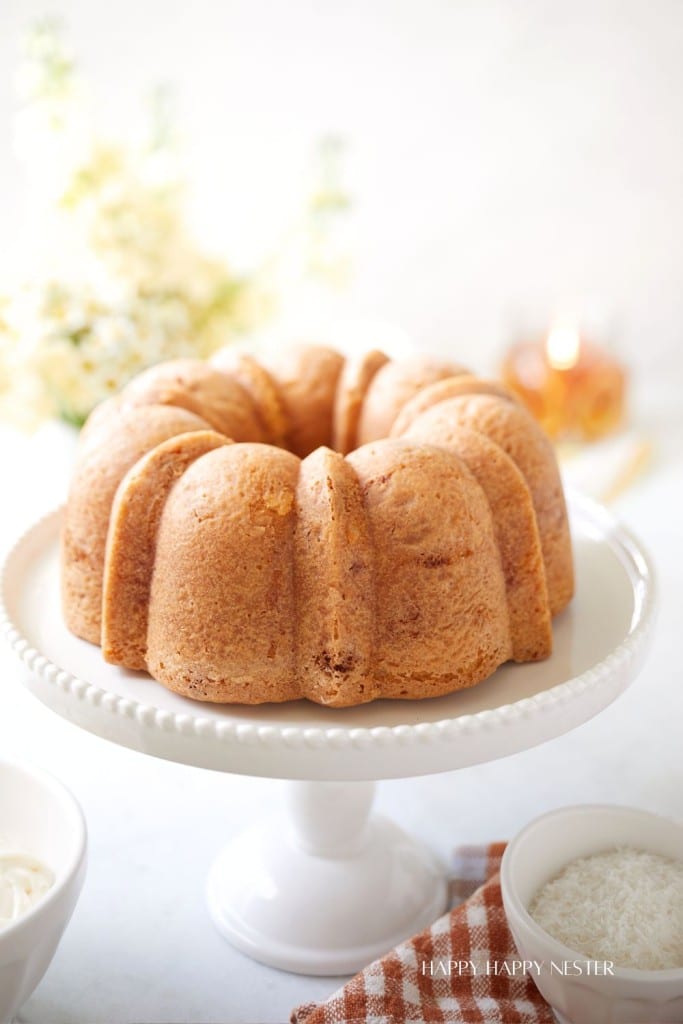 A golden-brown bundt cake displayed on a white pedestal cake stand, adorned with a fluted design. The background is softly blurred with light floral elements, and there are small bowls of white icing and coconut on the side, along with an orange-striped cloth.