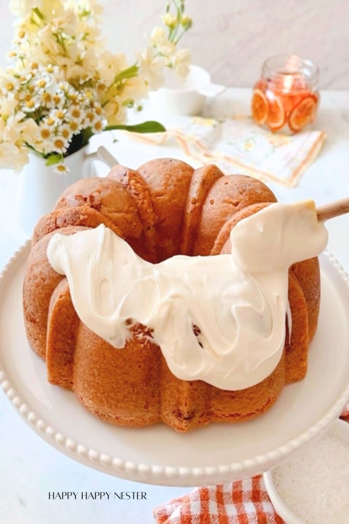 A freshly baked bundt cake is being frosted with a thick layer of cream. The cake sits on a white cake stand and is decorated with flowers in the background. A jar of dried orange slices and a checkered red and white cloth are also visible on the counter.