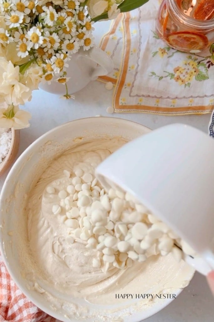 A bowl of cake batter is on a countertop. White chocolate chips are being poured into the batter from a separate bowl. Nearby are a bunch of daisies, a cup with floral patterns, and a napkin. The countertop also has a jar of honey and a container of powdered sugar.