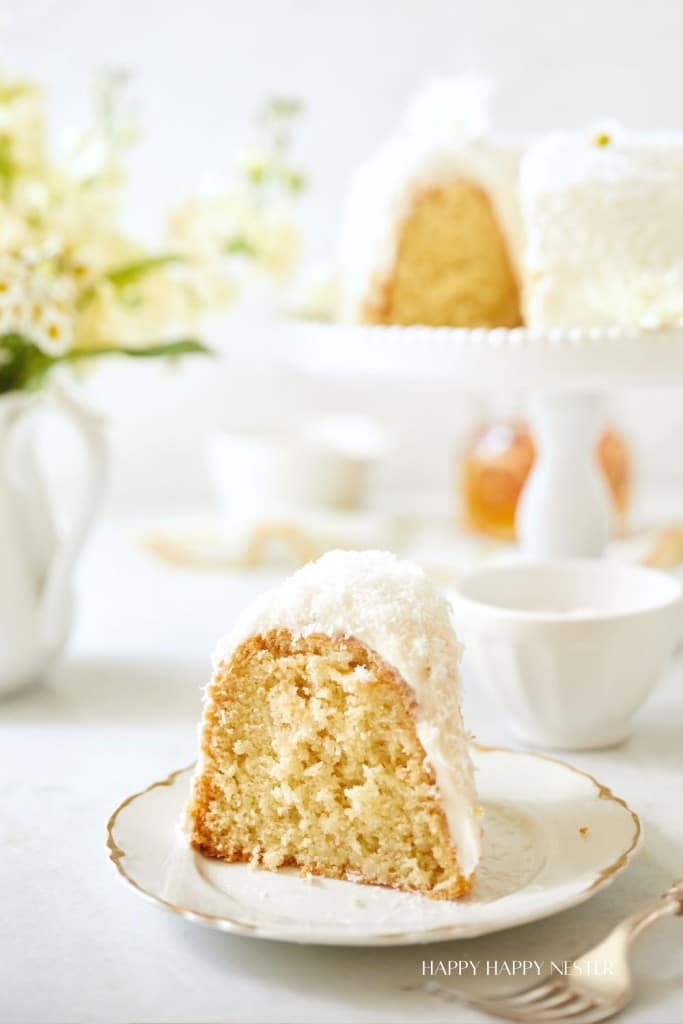 A slice of bundt cake with white icing and coconut flakes sits on a decorative plate. Behind it, more cake is displayed on a raised cake stand, and there are white flowers and tea cups in the background, creating a light and elegant setting.