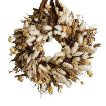 A rustic wreath is made from a mixture of dried grasses and cotton, creating a natural and earthy decoration. Hues of beige, brown, and white are prominent, and the wreath is suspended by a brown ribbon.