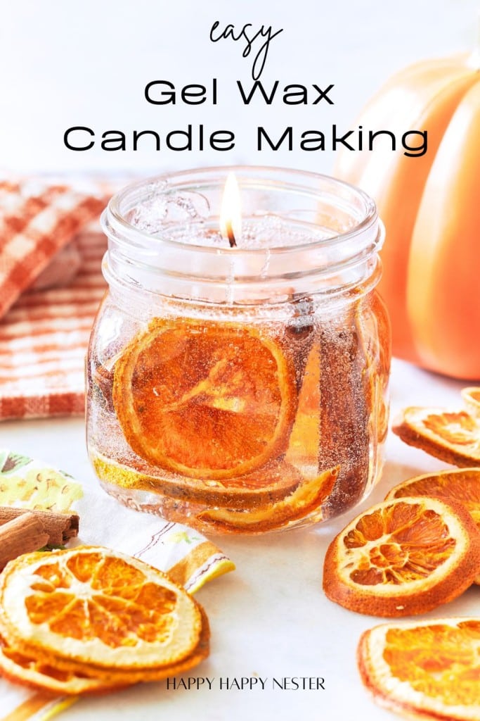 An image showcasing a lit candle in a glass jar with dried orange slices embedded in the gel wax. The text reads "Easy Gel Wax Candle Making." There are additional dried orange slices and a striped cloth in the background. The source is noted as "Happy Happy Nester.