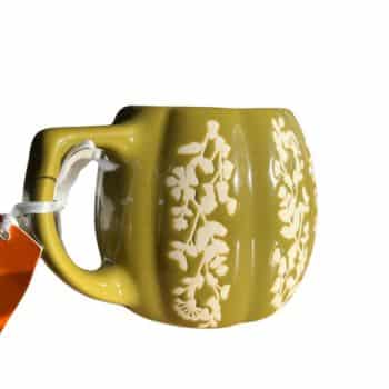 A round, yellow-green ceramic mug with embossed, white floral designs. The mug has a large handle and a white string with an orange tag attached to it. The mug is set against a plain white background.