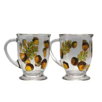 Two clear glass mugs with handles, each featuring a decorative pattern of acorns and oak leaves painted in autumn colors, primarily brown, green, and yellow. The mugs have a slightly flared base and are displayed side by side against a plain background.