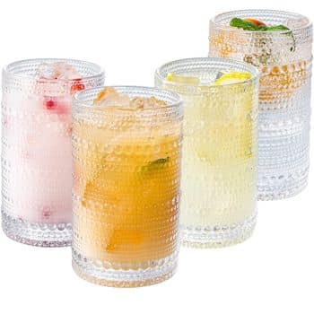 Four textured glass tumblers filled with various colorful beverages, including a pink drink with ice and strawberries, a brown drink with ice cubes, a pale yellow drink with ice and lemon slices, and a clear drink with ice and a mint garnish.