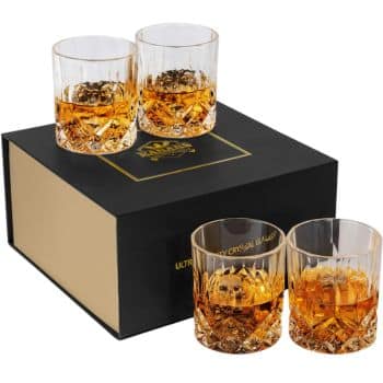 A set of four filled whiskey glasses positioned atop and in front of a black and gold box. The glasses feature intricate crystal designs, showcasing the amber liquid inside. The box lid displays a gold logo and the text "Ultra Fancy Crystal Glasses.