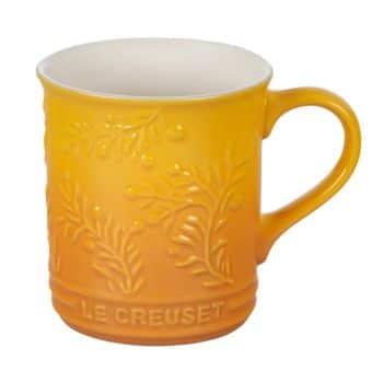 A yellow-orange Le Creuset ceramic mug featuring a textured, embossed design with branches and small berries. The mug has a smooth interior and handle, with the brand name "Le Creuset" engraved at the bottom.