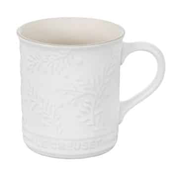 A white ceramic mug with a handle, featuring an embossed botanical pattern. "Le Creuset" is engraved at the bottom of the mug. The interior is a lighter shade of white.