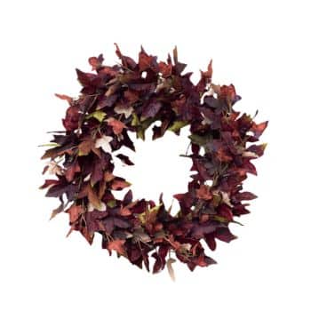 A circular autumn wreath made of artificial leaves in various shades of red, orange, and brown. The leaves are arranged densely, creating a full and vibrant appearance, symbolizing the fall season. The wreath is set against a plain white background.