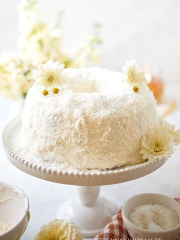 A beautifully crafted white cake with a fluffy texture, generously covered in shredded coconut, sits atop an elegant white cake stand. The cake is adorned with white flowers, and a small bowl of coconut shreds sits nearby on a checkered cloth. White flowers are in the background.