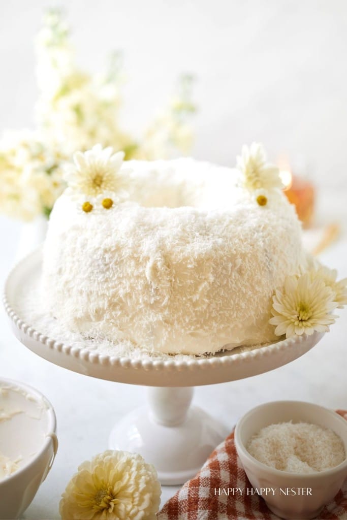 A beautifully crafted white cake with a fluffy texture, generously covered in shredded coconut, sits atop an elegant white cake stand. The cake is adorned with white flowers, and a small bowl of coconut shreds sits nearby on a checkered cloth. White flowers are in the background.