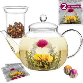 A clear glass teapot with blooming tea flowers inside, alongside a glass infuser. The tea flowers have yellow and pink blooms. A packet labeled "2 Bonus Tea Flowers Included" and two sealed tea flower packages are also visible.