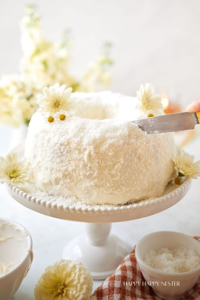 A white coconut cake is placed on a white cake stand. The cake is decorated with fluffy shredded coconut and white flowers. A hand is visible, holding a knife and frosting the side of the cake. A bowl and a napkin are nearby in the foreground.