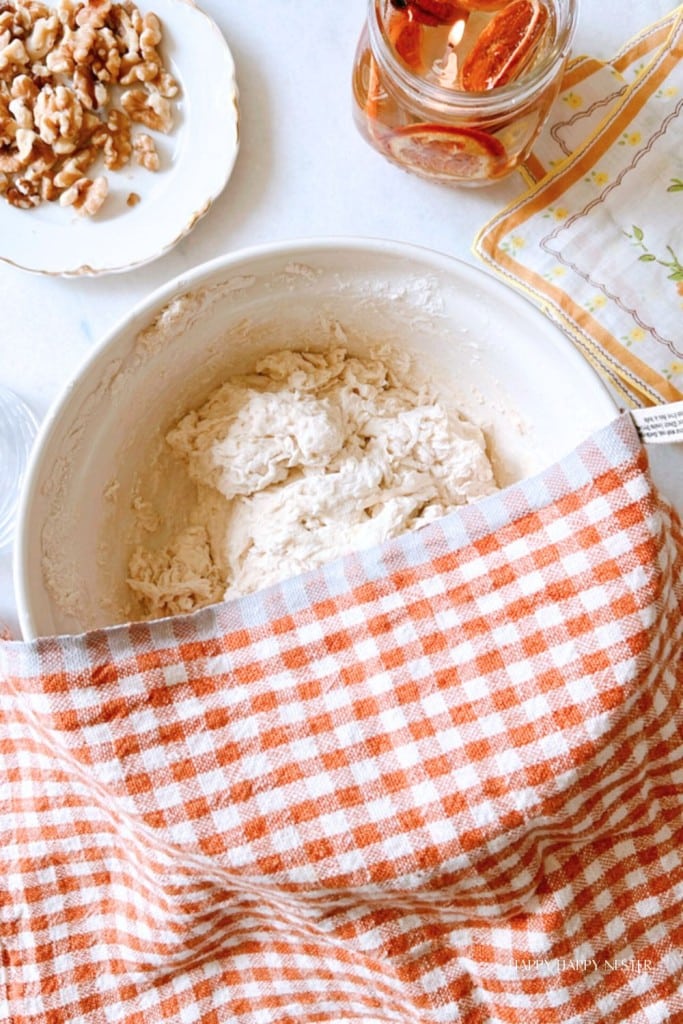 A bowl of dough covered with an orange and white checkered cloth sits on a surface next to a plate of chopped walnuts and an open jar of honey. A yellow-bordered napkin with floral patterns is placed beside the bowl.