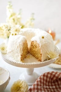 A frosted Bundt cake topped with shredded coconut is displayed on a white cake stand. A slice is cut from the cake, revealing its moist interior. The scene includes white flowers, a lit candle in the background, and a checked napkin in the foreground.