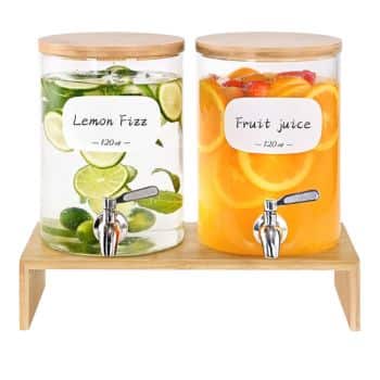 Two glass beverage dispensers with wooden lids sit on a wooden stand. The left dispenser is labeled "Lemon Fizz" and contains lime slices and mint leaves. The right dispenser is labeled "Fruit Juice" and contains orange and lemon slices. Each has a metal spout.