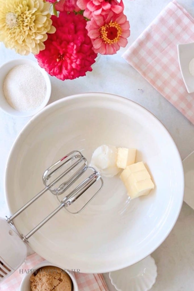 A white bowl containing butter and shortening is placed on a marble countertop. An electric mixer is positioned over the bowl. Surrounding the bowl are ingredients, such as sugar and flour, and a pink and white checkered cloth. Bright flowers are in the top left.