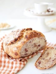 A rustic loaf of freshly baked bread with fruit and nuts, resting on an orange and white checkered cloth. One slice is cut and placed nearby. In the background, a white cup and saucer are visible on a white cake stand.