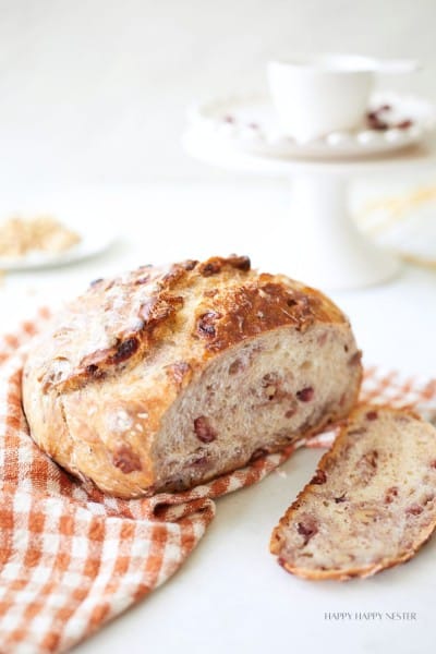 A rustic loaf of freshly baked bread with fruit and nuts, resting on an orange and white checkered cloth. One slice is cut and placed nearby. In the background, a white cup and saucer are visible on a white cake stand.