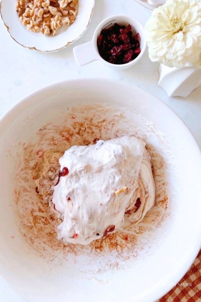 A large white mixing bowl containing partially mixed dough with visible cranberry bits. Next to the bowl is a small dish of cranberries and another dish of walnuts. A vase with a white flower is placed to the side on the countertop.