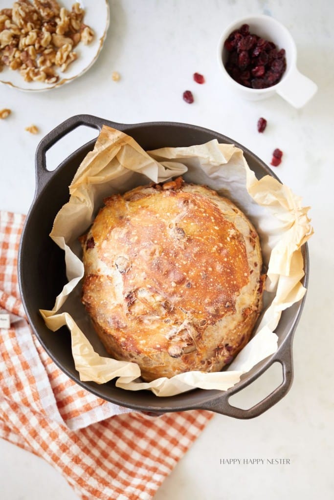 A round loaf of bread with a golden crust sits in a cast iron pot lined with parchment paper. Nearby, a small bowl of walnuts, a white cup of dried cranberries, and a checkered orange and white kitchen towel are visible on a white surface.