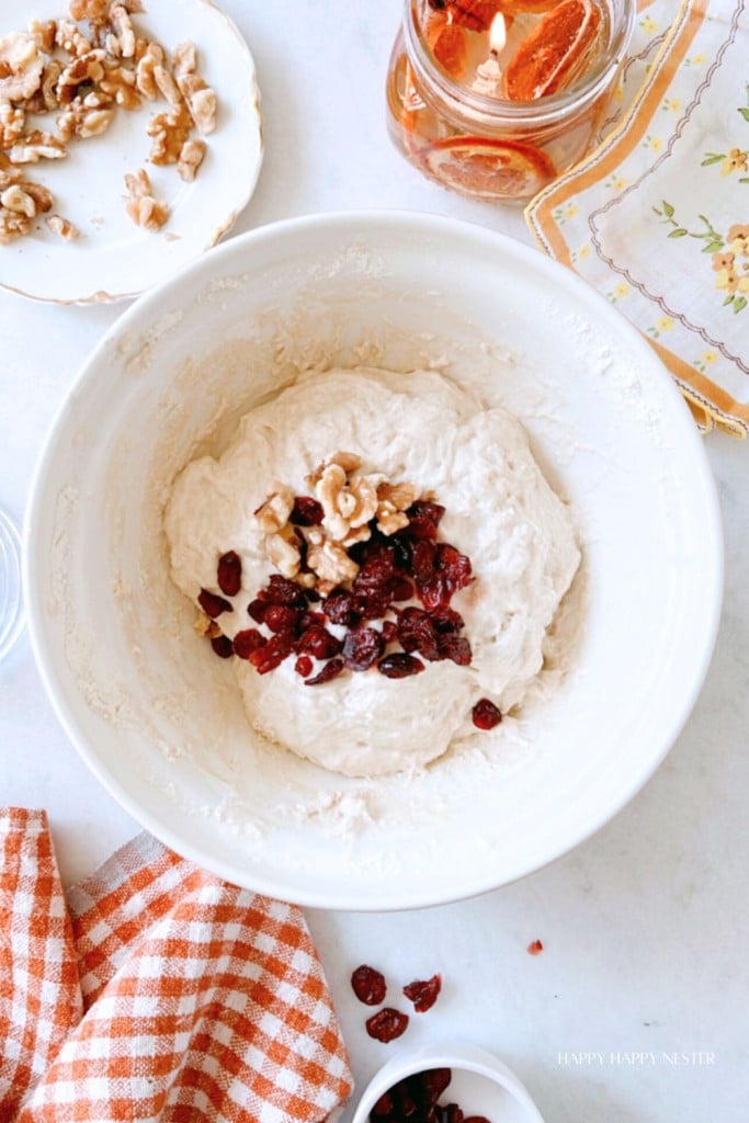 A bowl contains bread dough with added dried cranberries and chopped walnuts. Surrounding it are a plate of walnuts, a lit candle, an orange-checked cloth, and a small floral dish.