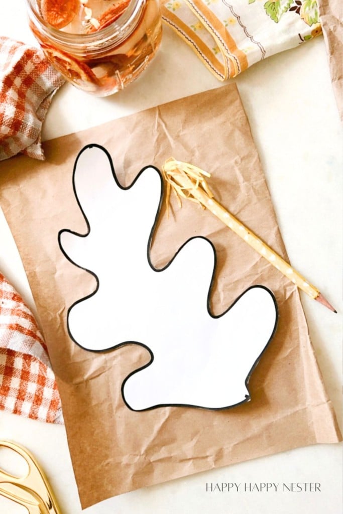 A shaped piece of white paper with a black outline, resembling an oak leaf, lies on a brown paper sheet. A gold pen with a white tip rests beside it. Surrounding the paper are fabric pieces in fall patterns, a pair of scissors, and a glass jar with dried orange slices.