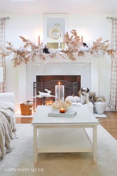 A cozy living room with a white coffee table, decorated with candles and seashells. A dog sits near a fireplace adorned with large, white leaf garlands and red candles. The room is softly lit and has neutral-toned furniture and decor.