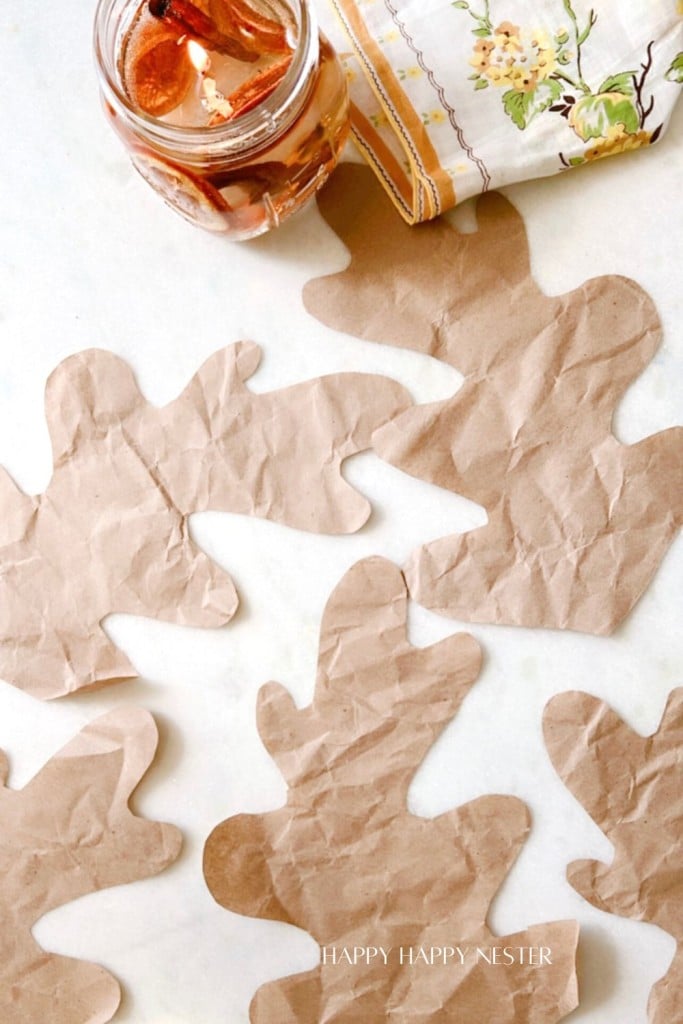 A flat lay photo shows several crumpled, brown paper cut-outs in the shape of oak leaves scattered on a white surface. A jar with cinnamon sticks and light-colored fabric with floral designs is visible in the upper corner. Text reads "HAPPY HAPPY NESTER.