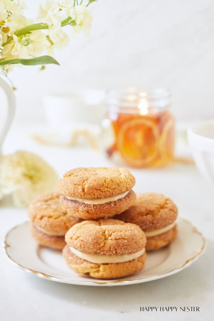 A white plate holds a stack of three sandwich cookies with golden-brown shells and a creamy filling. Behind them is a jar with sliced fruits, part of a candle. The background includes white flowers in a vase and blurred kitchen items. The scene is bright and inviting.