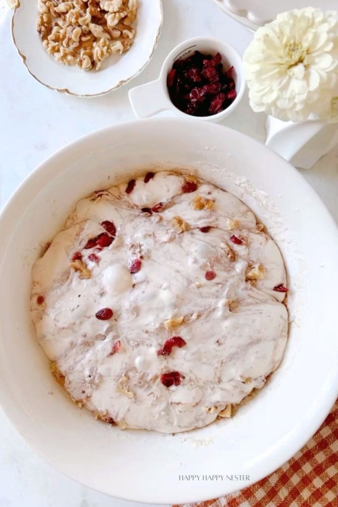 A bowl mixed with a creamy white batter containing visible chunks of walnuts and cranberries. Surrounding the bowl are a plate of walnuts, a small white dish of cranberries, and a white flower in a vase. A "HAPPY HAPPY NESTER" watermark is at the bottom.