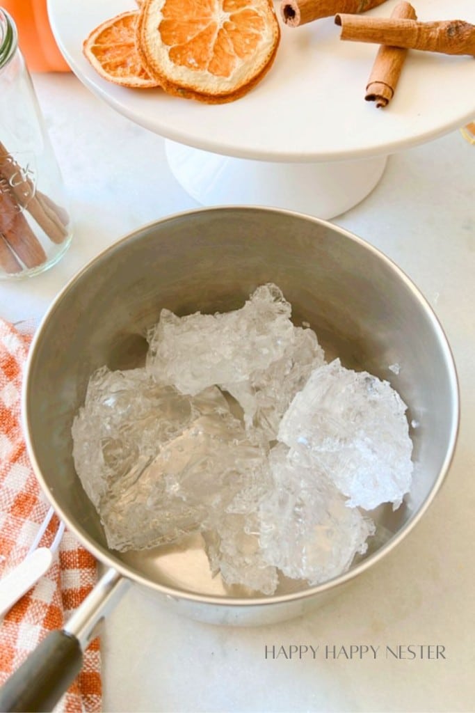 A metal bowl filled with large ice chunks sits on a countertop next to a red and white striped cloth. In the background, dried orange slices and cinnamon sticks are displayed on a white cake stand. Text at the bottom reads "Happy Happy Nester.