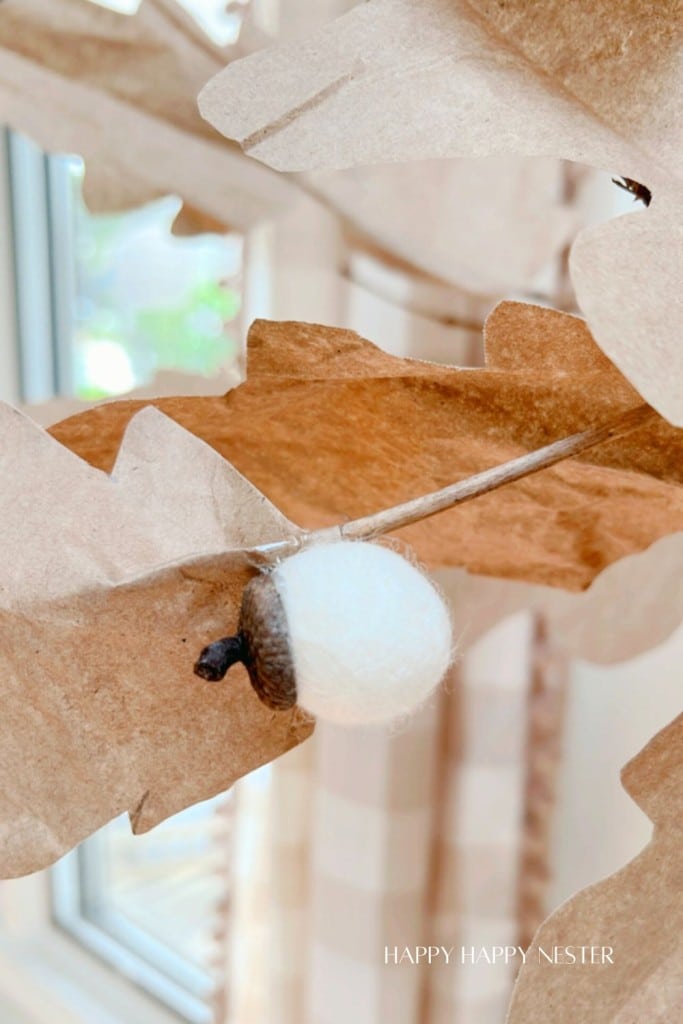 Close-up of a craft displaying a felted acorn attached to a faux autumn leaf made of brown paper. The background shows a window with blurred outdoor light coming through. A faint "HAPPY HAPPY NESTER" watermark is visible at the bottom right corner.