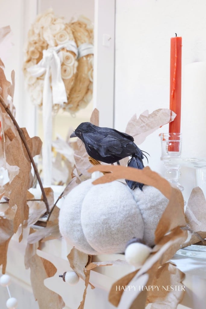 A black artificial bird perched on a white, fabric pumpkin amidst dried leaves and branches. Behind it, a red candle and a decorative wreath with a ribbon are visible, set against a light-colored background. The words "HAPPY HAPPY NESTER" are at the bottom.