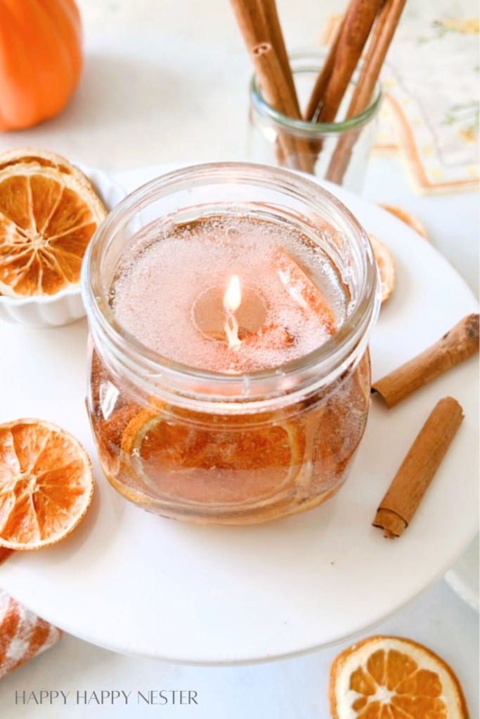 A clear jar candle filled with a translucent, orange-colored wax and decorated with dried orange slices is lit on a white surface. Cinnamon sticks are placed beside the jar and in a glass container in the background. Dried orange slices also adorn the table.