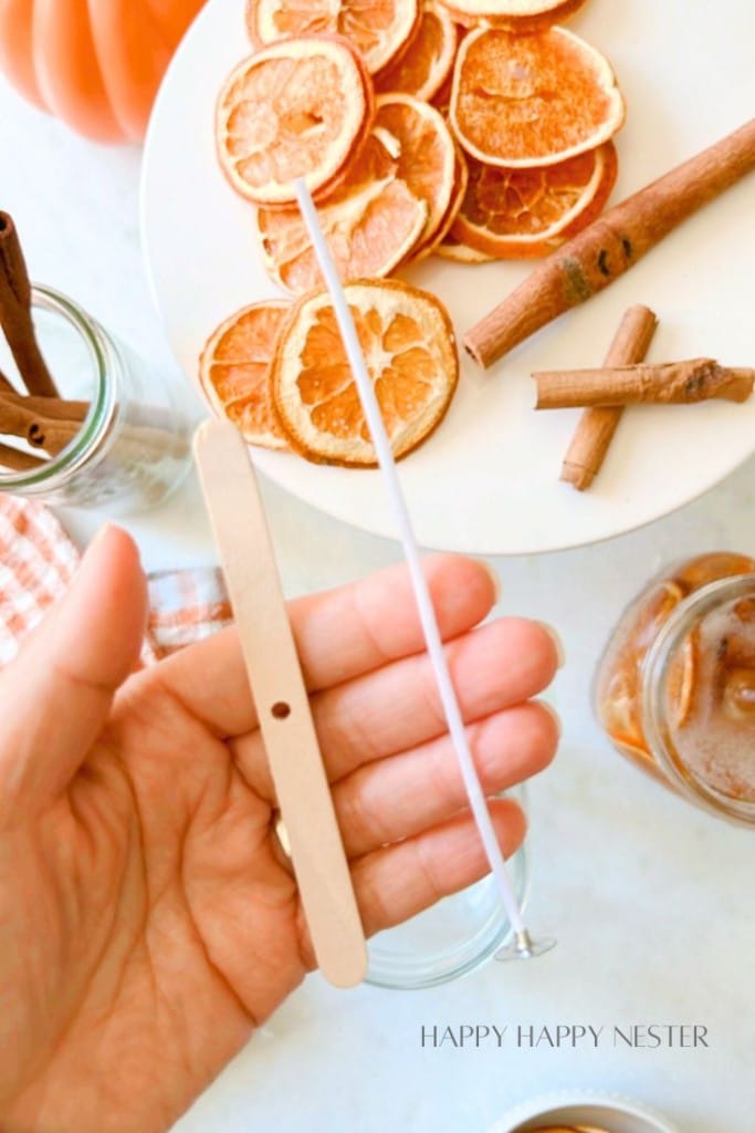 A hand holding a wooden candle wick holder and a metal wick. On the table, there is a plate of dried orange slices, a couple of glass jars with cinnamon sticks, and a cup of iced coffee. A pumpkin and a cloth napkin are also visible in the background.