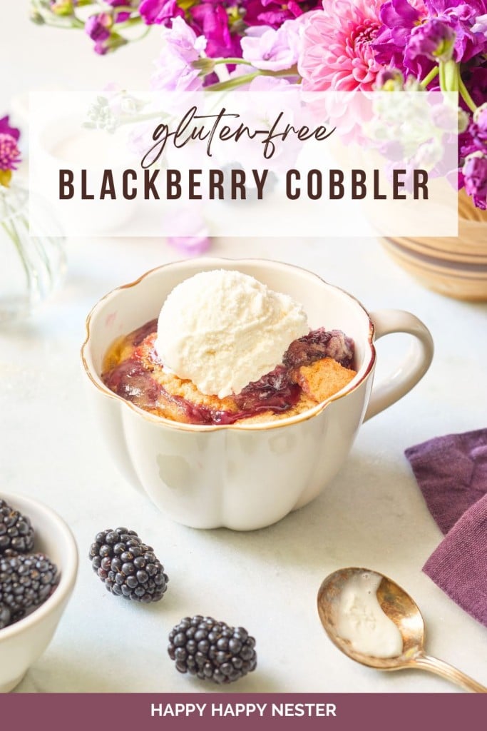 A ceramic mug filled with gluten-free blackberry cobbler, topped with a scoop of vanilla ice cream. Fresh blackberries, a spoon, and a napkin sit nearby. The background includes a bouquet of purple and pink flowers. The text overlay reads "Gluten-Free Blackberry Cobbler.