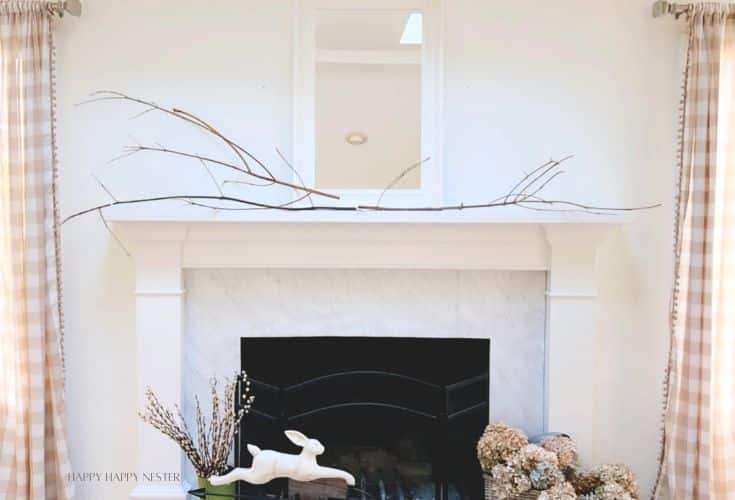 A fireplace mantle adorned with long, thin branches, a white ceramic rabbit figurine, and dried floral arrangements. The wall and mantle are white, and there are beige and white checkered curtains framing the scene. A mirror is mounted above the mantle.