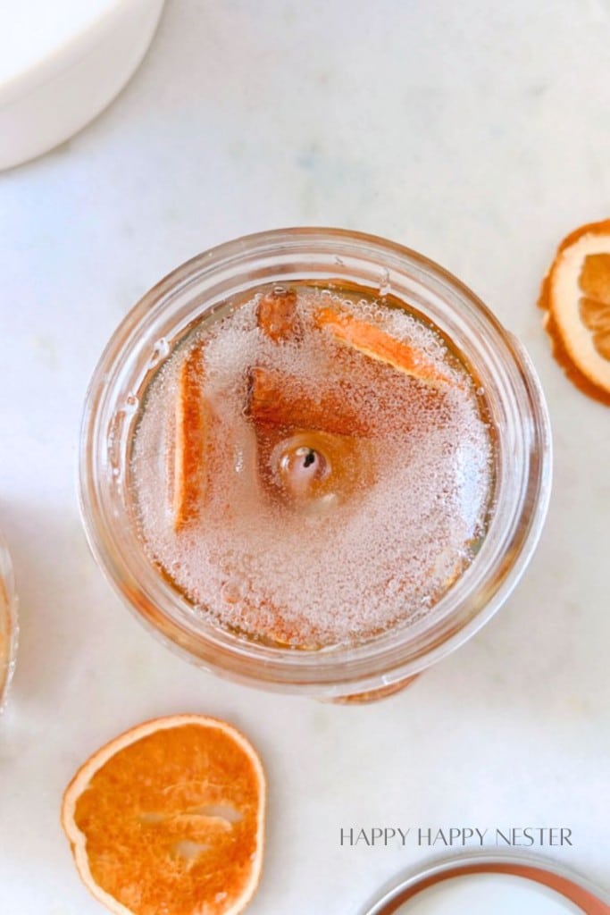 Overhead view of a glass jar filled with a bubbly beverage and garnished with orange slices. Dried orange slices are scattered on the surface around the jar. The text "HAPPY HAPPY NESTER" is faintly visible at the bottom right corner.