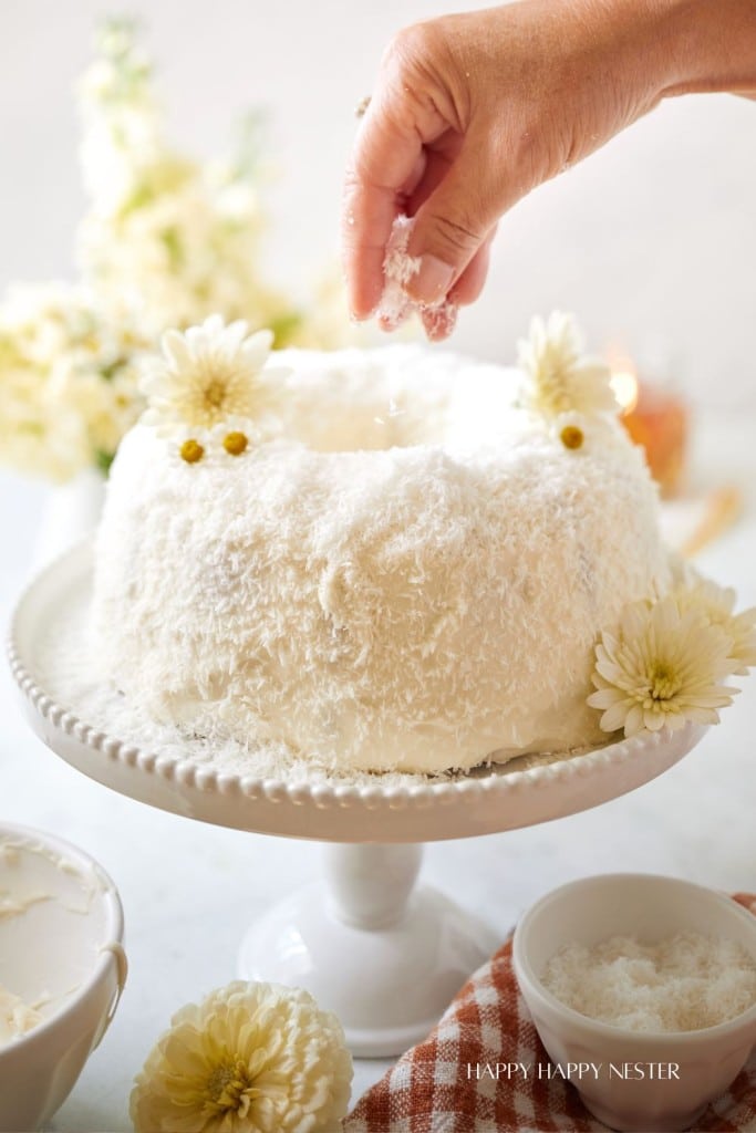 A hand sprinkles shredded coconut onto a white frosted cake on a pedestal stand. The cake is garnished with white flowers and surrounded by a bowl of frosting, a small dish of coconut, and a checkered cloth. In the background, there are cream-colored flowers.