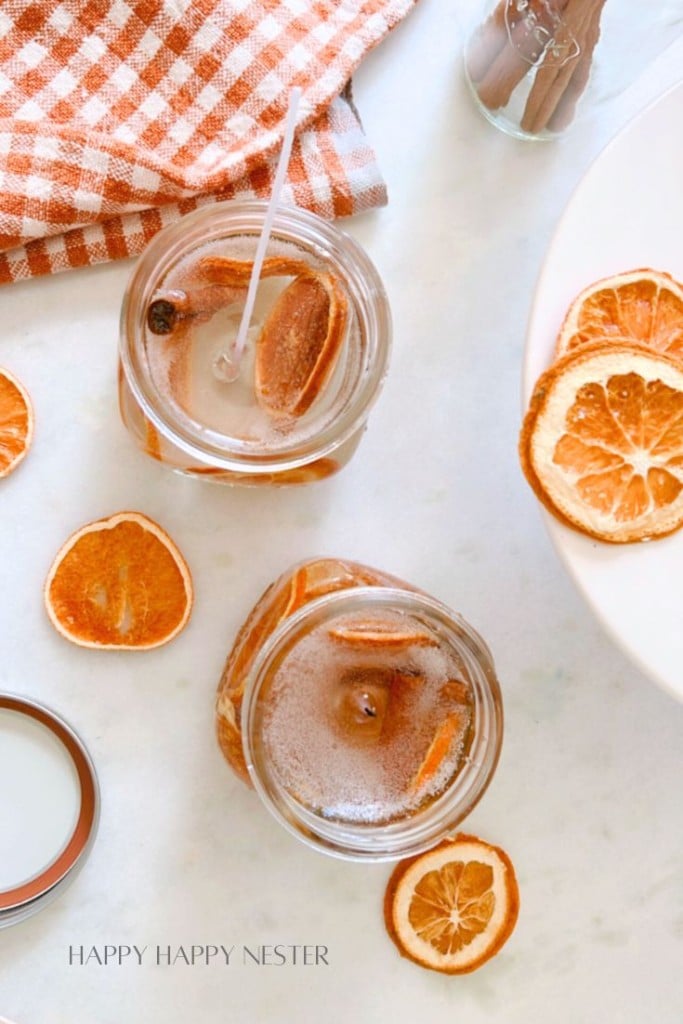 Two mason jars filled with a light-colored beverage, garnished with dried orange slices, and stirring sticks. The jars are placed on a light surface surrounded by additional dried orange slices. An orange and white checkered cloth is placed in the upper left corner.