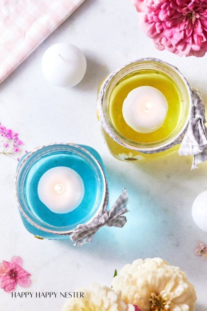 Overhead view of a blue and yellow candle in glass jars, each with a lit tealight inside. The jars are decorated with gray checkered ribbon bows. Surrounding the candles are white and pink flowers, and white round candles on a white surface.