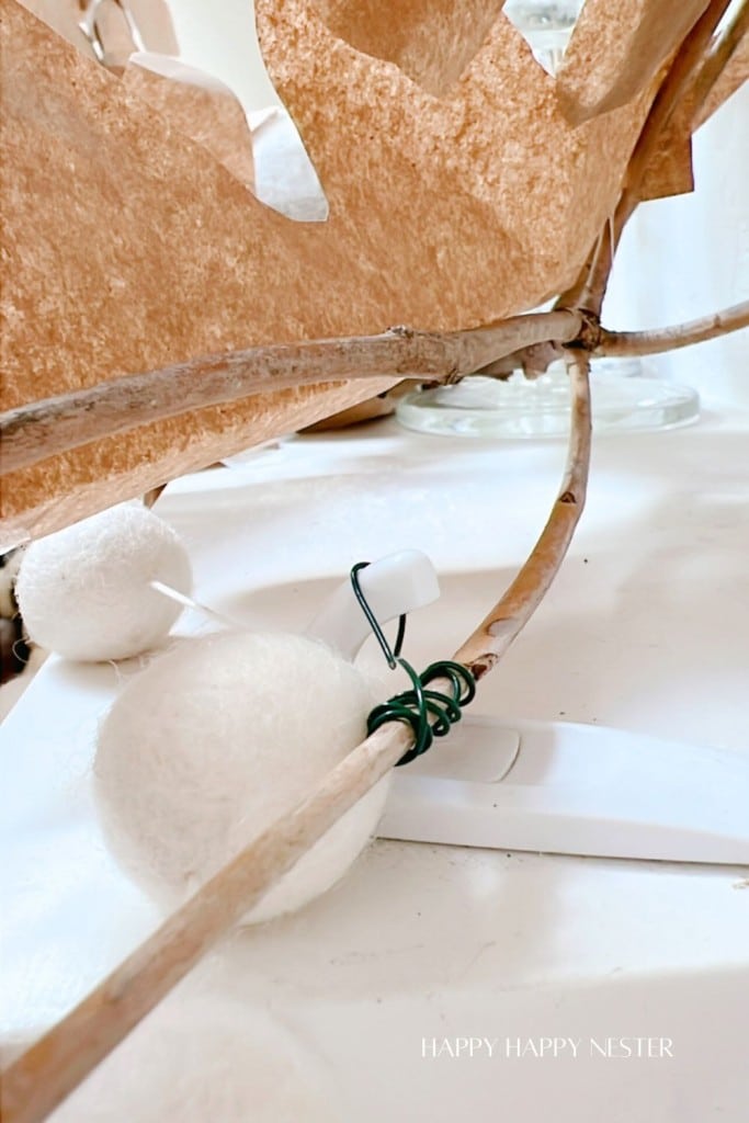 Close-up of a decorative setup featuring white felt balls attached to brown branches with green wire. A paper-like brown material forms a backdrop. The image has a soft, cozy aesthetic. The words “HAPPY HAPPY NESTER” are visible at the bottom right corner.