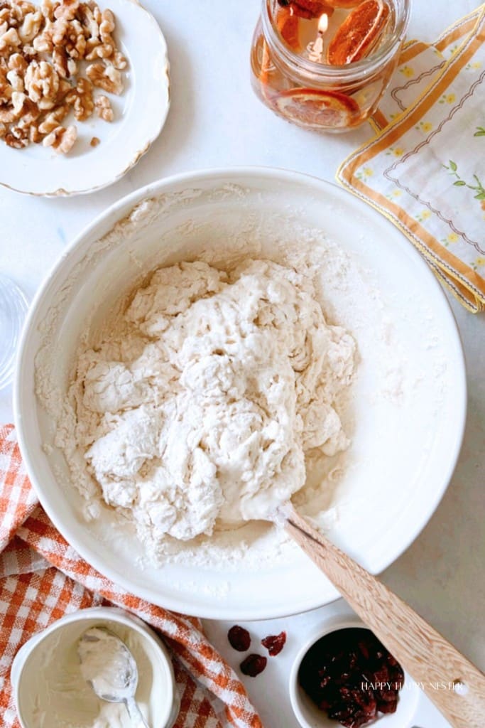 An overhead view of a mixing bowl containing dough being stirred with a wooden spoon. Surrounding the bowl are ingredients like nuts, dried cranberries, a small dish of cream, a checkered cloth, and a glass of amber-colored liquid on a patterned cloth.