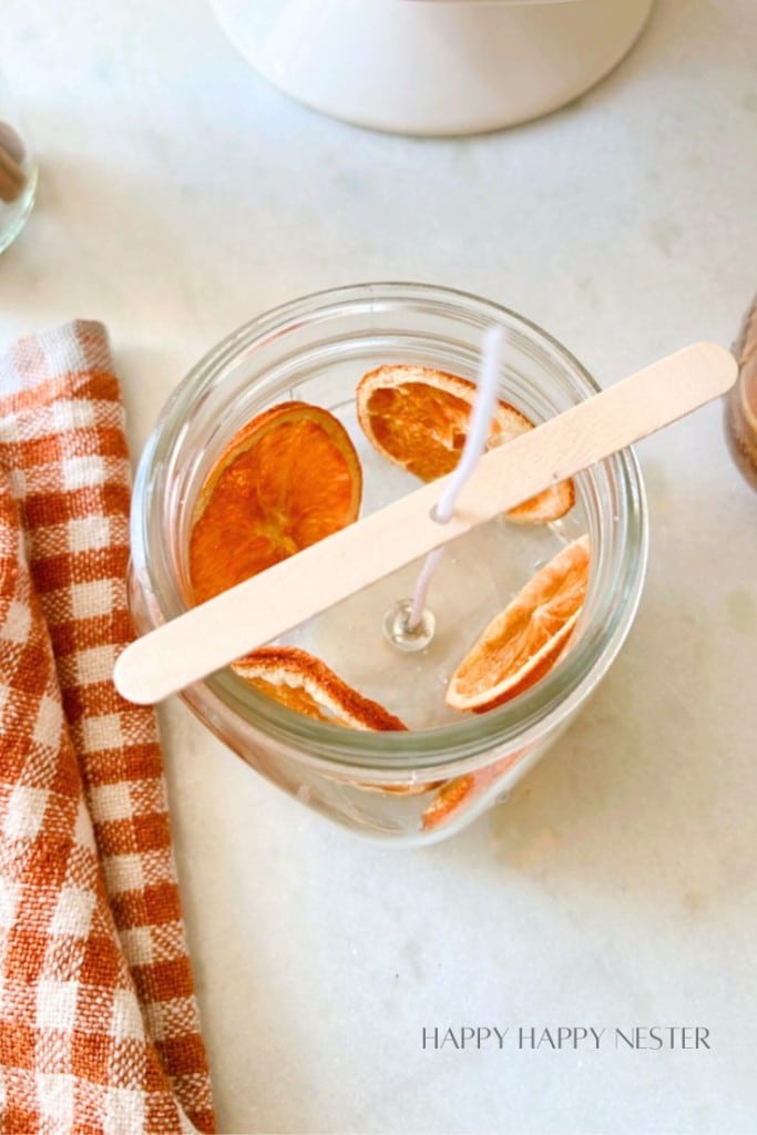 A glass jar with a wooden stick placed across its top, holding a wick in the center of the jar. The jar is filled with clear liquid and adorned with slices of orange. A gingham-patterned cloth lies beside the jar, and text at the bottom reads "HAPPY HAPPY NESTER.