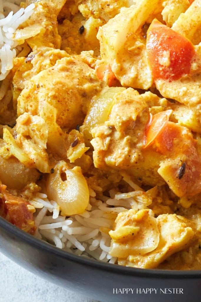 A close-up image of a dish consisting of rice topped with a creamy curry. The curry contains chunks of vegetables including tomatoes and onions, and appears to have a yellowish-orange hue, indicating the presence of spices. The dish is in a black bowl.