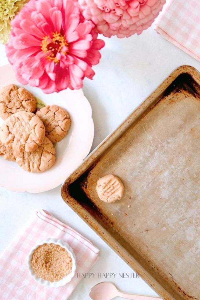 A baking tray with one peanut butter cookie on it is placed next to a plate of more cookies, a bowl of brown sugar, and a spoon. There are pink flowers and patterned napkins in the background. The words "Happy Happy Nester" are visible at the bottom of the image.