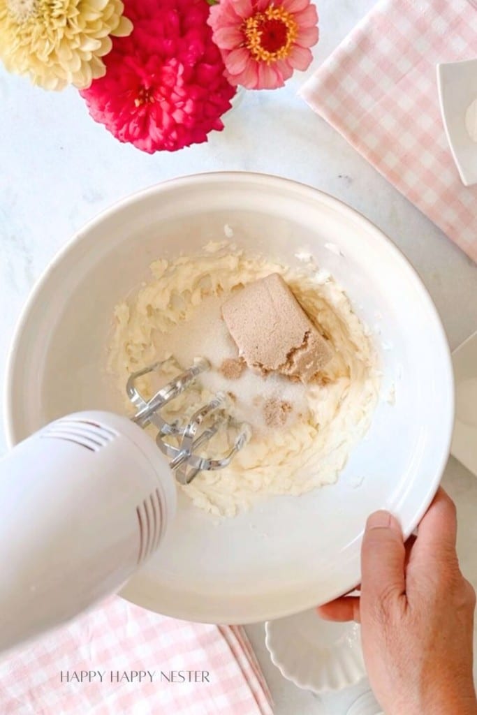 A hand holding a mixer is blending ingredients in a white bowl. The bowl contains cream, brown sugar, and other ingredients. The scene is set on a marble surface with pink checkered cloth and bright flowers in the background, creating a cozy, homey atmosphere.