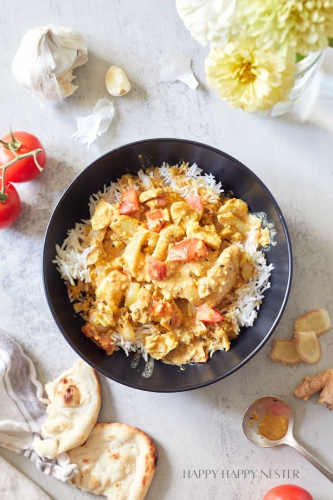 A bowl of rice topped with a creamy chicken curry garnished with diced tomatoes. Surrounding the bowl are tomatoes, garlic cloves, a piece of naan, fresh ginger, a small bowl of spices, and a striped linen. Yellow flowers in a vase are also visible in the corner.